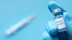 Hand wearing a medical glove holding a vial labled "COVID-19 vaccine," a syringe is out of focus in the background