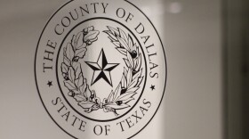 A transparent version of the Dallas County seal on a glass door. The seal includes the words "The County of Dallas - State of Texas" as well as a star in the center of two curved branches of leaves.