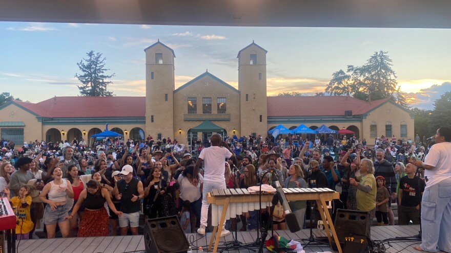 A man in a white tee, white pants and sneakers looks out at a packed crowd of people from a stage. Behind him is a xylophone, a sound monitor, and other band equipment. Behind the packed audience are some blue umbrellas and the Denver City Park Pavilion, a tall tan building with two prominent towers. The sky is a soft sunset. 
