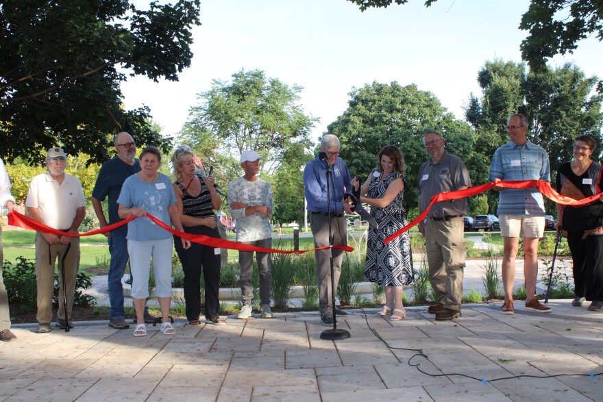 A group of people stand behind a red ribbon with a big pair of scissors, cutting the ribbon. 