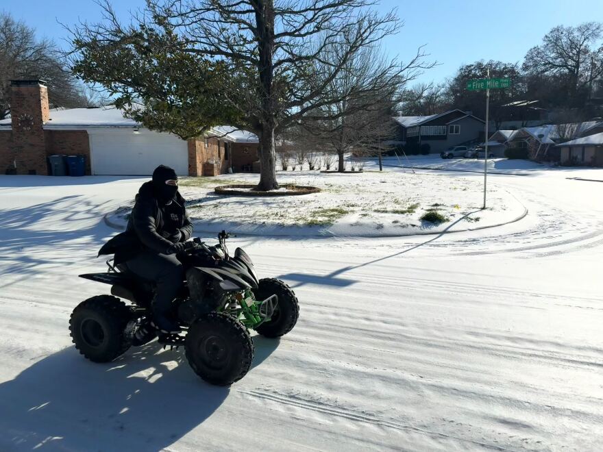 A person in a balaclava on a black and green four-wheeler drives across a snow-covered residential street. 