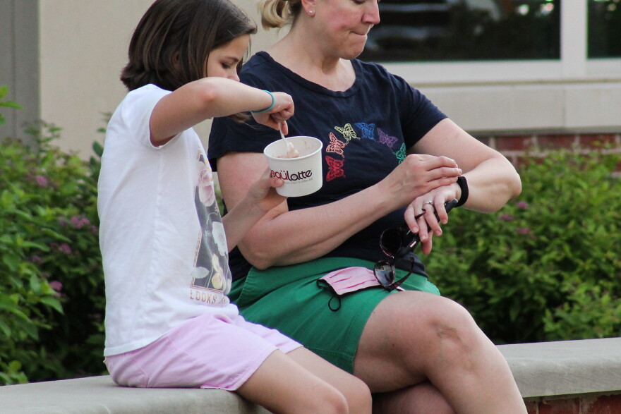 A child sits on a bench with an adult while eating a cup of ice cream. 