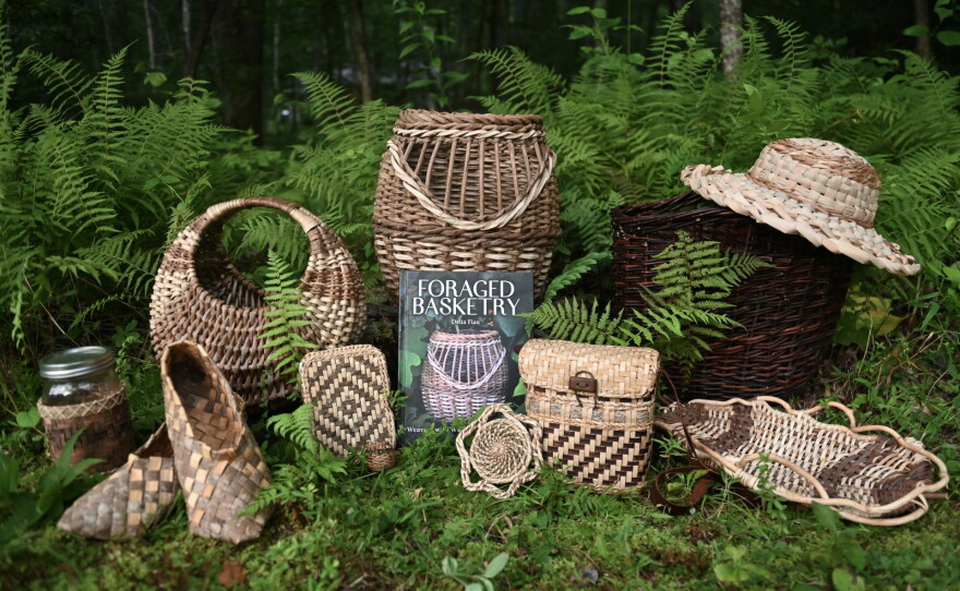 Foraged Basketry book is displayed among handmade woven baskets, a hat and other woven pieces in a wooded area surrounded by ferns.