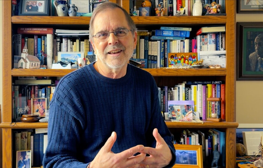 Speaking in front of a bookcase, a man smiles and weaves his fingers together
