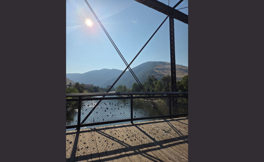 View from a footbridge overlooking a sunlit river, with tree-covered mountains rising in the distance beneath a clear blue sky.