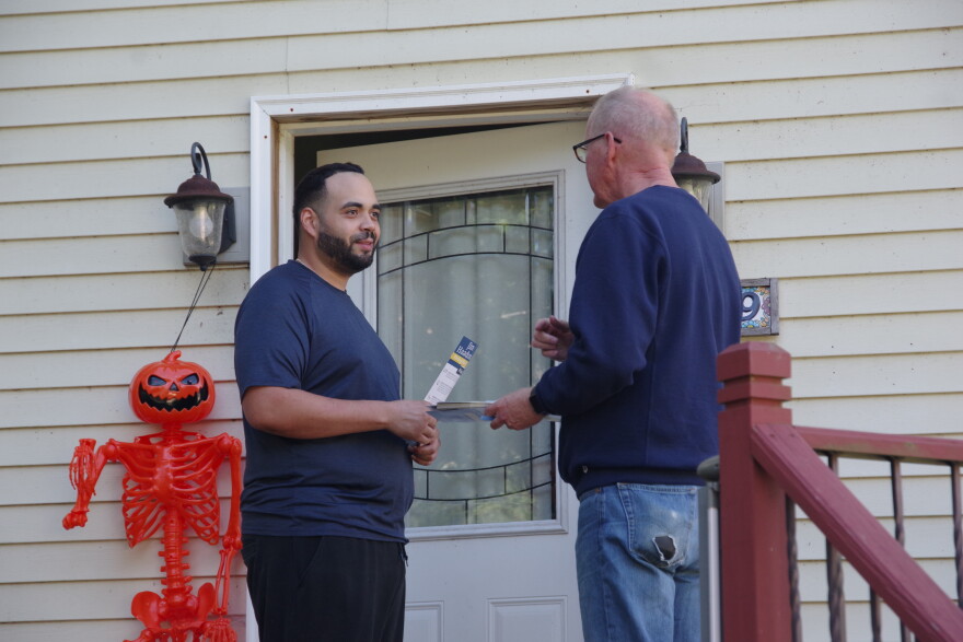 Two men talk in front of the white front door of a white wooden house. To the right of the door stands an orange skeleton with a jack-o-lantern. 