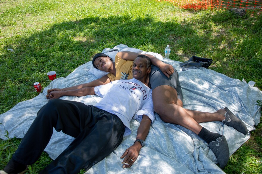 Two residents of Camp Rhonda lay on a grey blanket spread out under the shade of a tree. Demetria lies horizontally with Sam's head resting on her stomach while the pair hold hands.
