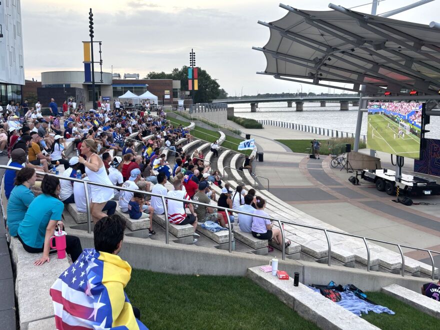 A crowd in a park-like amphitheater watch a screen on a summer evening. 