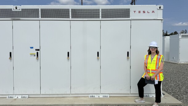 A woman in a bright yellow safety vest and white hard hat stands smiling alongside a row of vented white lockers that contain energy storage batteries.