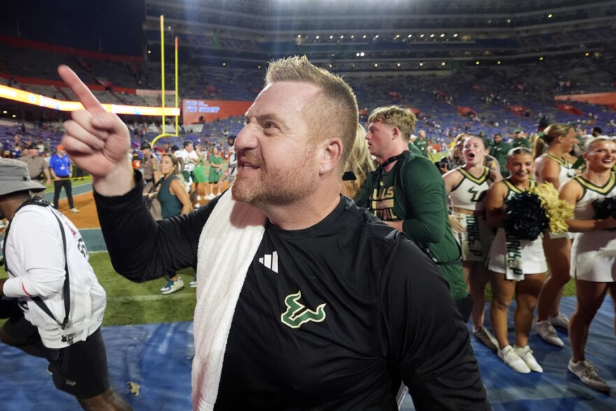 Man with short reddish hair wearing a long-sleeve black athletic shirt and pointing to the left with cheerleaders and a crowd behind him