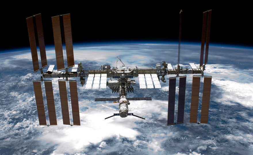 The Earth is seen from space with the ISS in front of it. The ISS features 16 long panels jutting out into space.