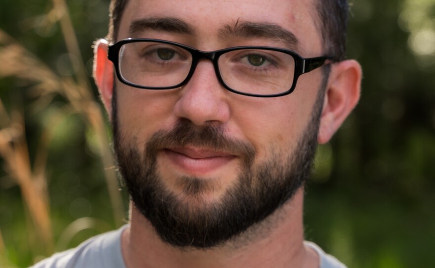 A close-up of Luke Myers wearing black-framed glasses and sporting a trimmed beard. He's slightly smiling with greenery in the background.