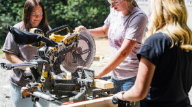 A young woman with a ponytail and clear work goggles uses a saw, with sawdust flecks flying in the air around her, as two other women stand watching. 