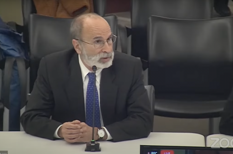 Mike Bromwich sits at a table with a mic in front of him, his hand crossed.