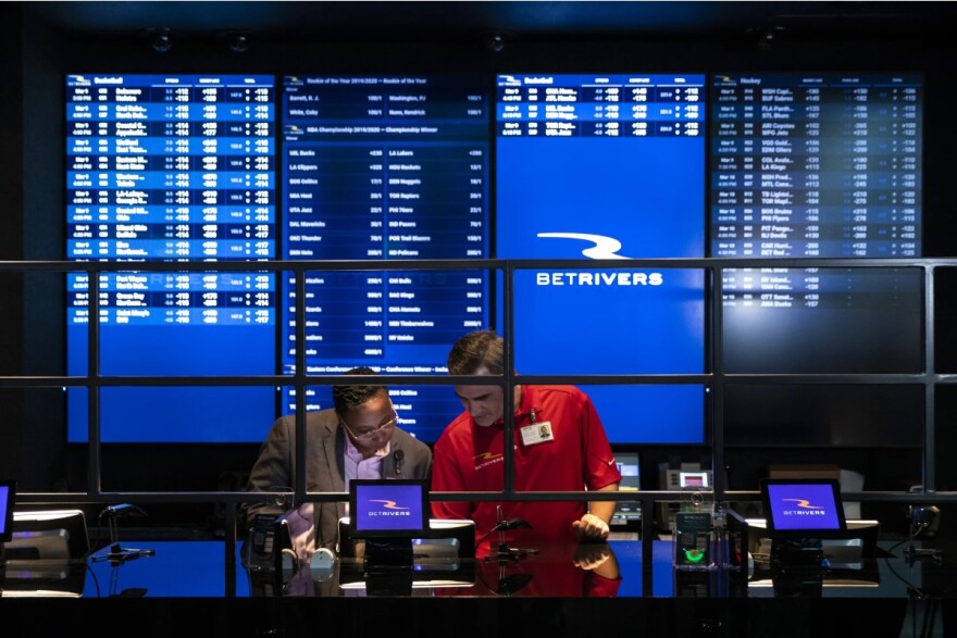 Two people are standing in front of large video boards at a brick-and-mortar sportsbook.