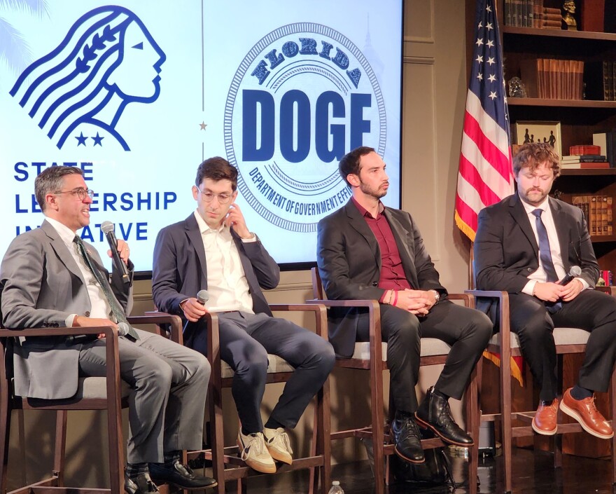 4 men in blazers sitting in tall chairs side by side having a discussion on a stage with a projected background that reads Florida DOGE 