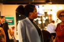 A Black woman in a black turtleneck and gray or light blue blazer stands in profile in a photo at an indoor election watch party. 