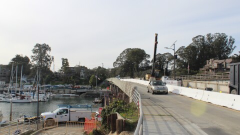 A car drives over a bridge that's under construction. Boats sit in a harbor to the left.