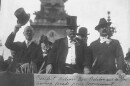 Historical black and white photograph from 1916 showing President Woodrow Wilson tipping his top hat alongside Indiana Governor Samuel M. Ralston and Indianapolis Mayor Joseph E. Bell as they view a parade from a reviewing stand in front of the Soldiers and Sailors Monument.