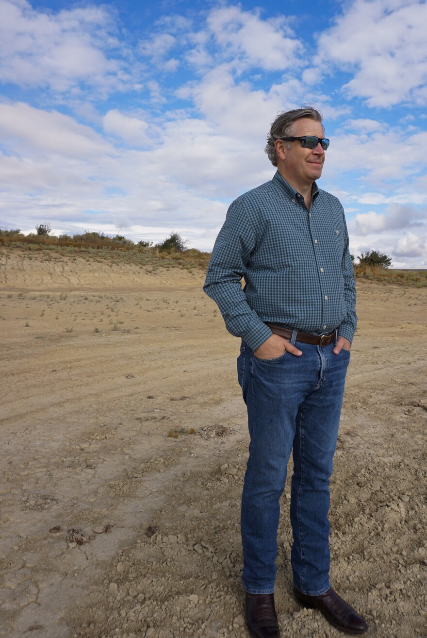A man wearing sun glasses, a blue plaid shirt and jeans gazes to the right, standing on a dirt lot.