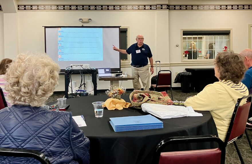 A man in a blue shirt points to a display as he gives a talk to older Vermonters about computer scams.