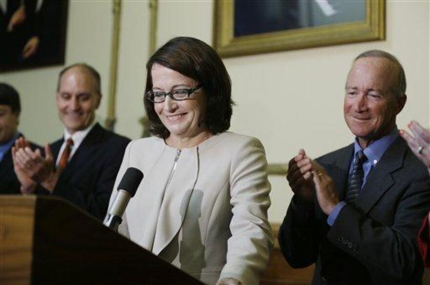 Loretta Rush stands smiling at a podium while Gov. Mitch Daniels, standing to her right, applauds during a formal ceremony.