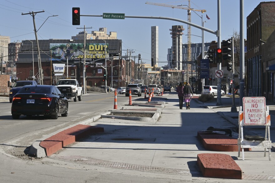 Exterior photo of a busy street at left. At right two people are walking away from the camera on a freshly poured concrete space.