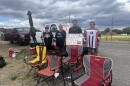 Four people hold protest signs and flags in front of a truck with more signs on it. Lawn chairs are set up in front of them.
