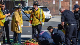 Firefighters in yellow jackets stand on the left while paramedics in blue attend to a person lying on the ground.