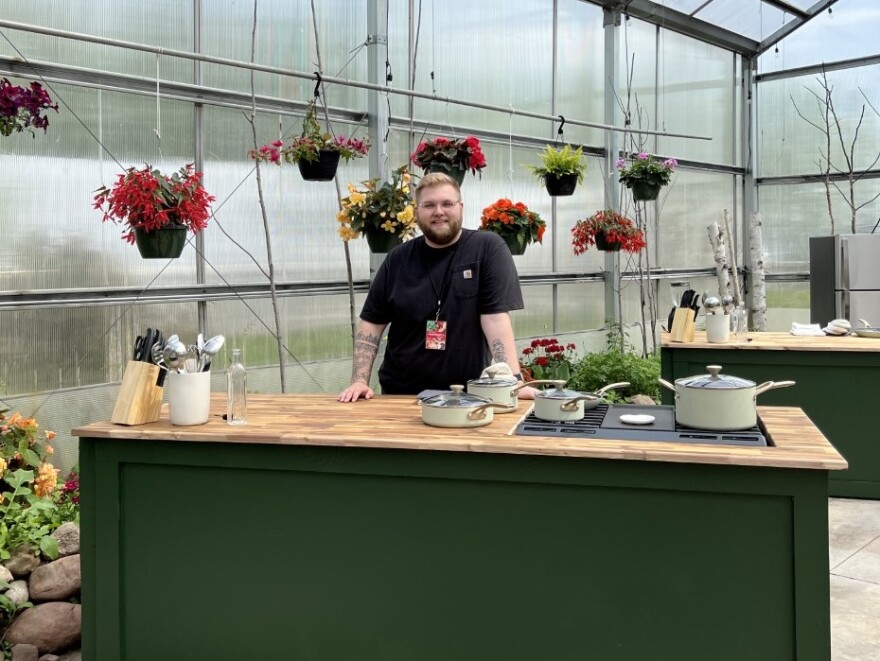 A person stands behind a kitchen island station in a greenhouse with plants hanging behind