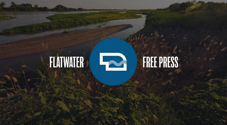 Flatwater Free Press logo set against a transparent backdrop of an aerial view of a marshy area