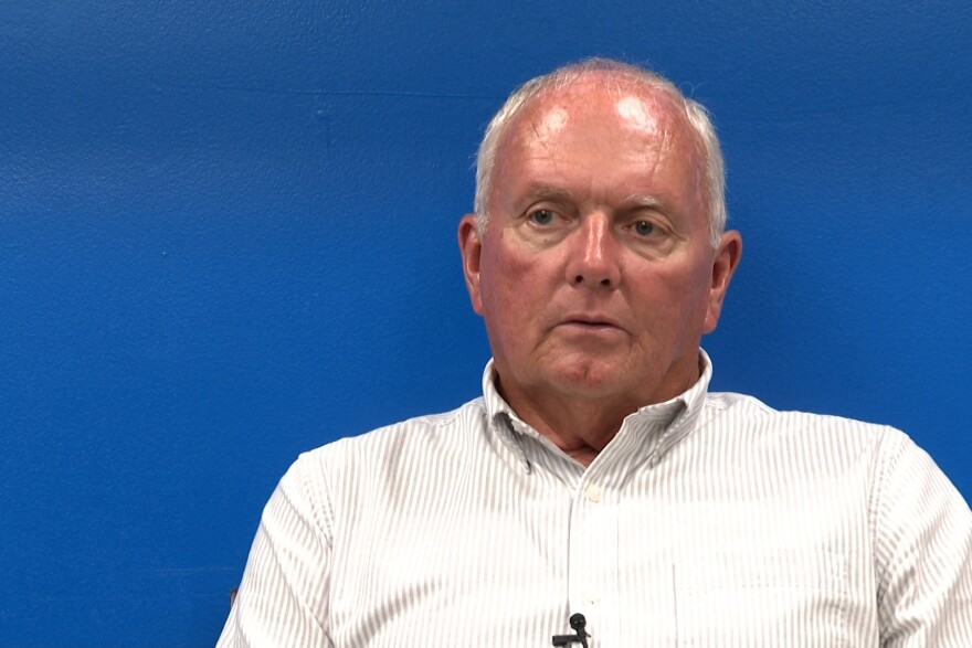 Town Manager Mike Farmer sits in front of a blue background. He is a white man with white hair wearing a white button down shirt.