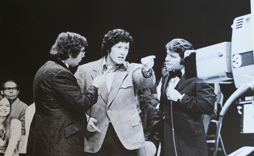  man in 1970s stands flanked by two other men and speaks into a television camera