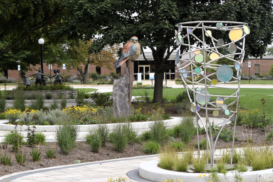 A garden features a large bird sculpture on a stone pedestal and a tall metal sculpture with colorful glass discs, surrounded by greenery and flowers, with a building in the background.