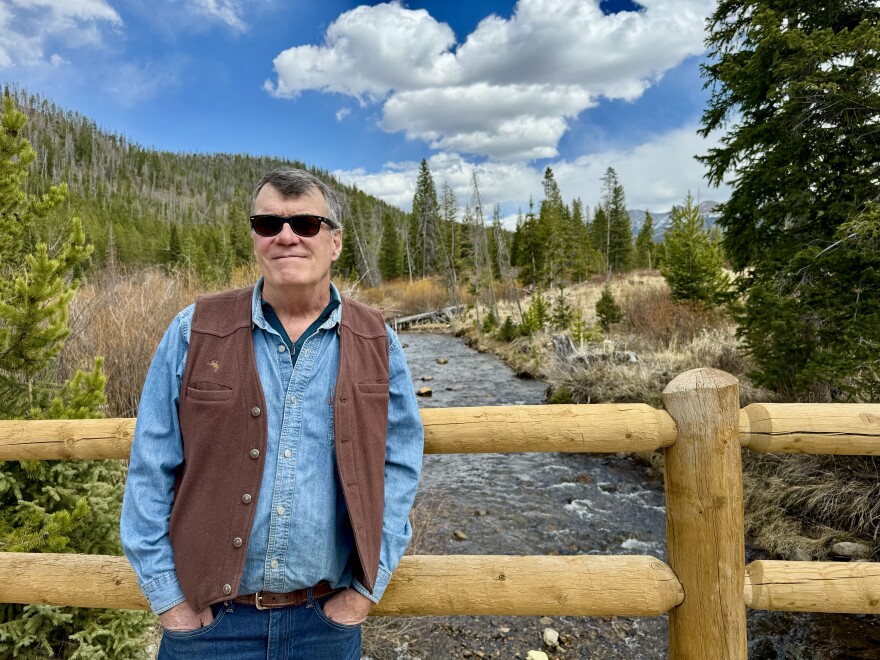 A man in a western vest poses on a bridge over a flowing creek