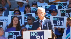 Denver District Attorney John Walsh speaks at a podium surrounded by people holding signs that say "No on 81"