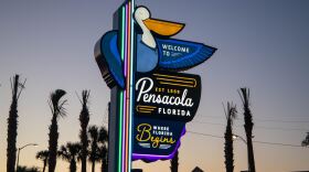 A brightly lit “Welcome to Pensacola, Florida” sign featuring a pelican and the slogan “Where Florida Begins,” illuminated by neon lights at dusk with palm trees silhouetted in the background.