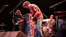 A man on stage plays a guitar while looking at the ground, with a bass player and a drummer out of focus in the background.