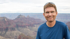 A man with brown hair wears a blue shirt and stands in front of a canyon. He smiles at the camera