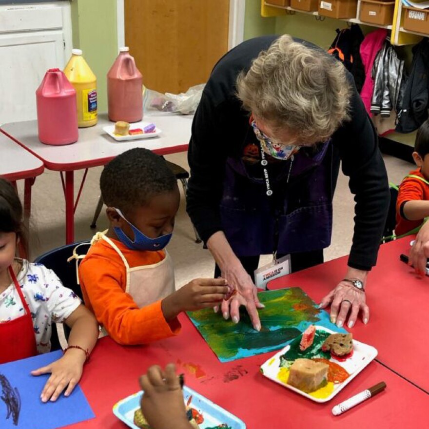 An older woman stands over a child, looking at the young person's painting.