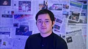 Emilio Milo wearing wire glasses and a black shirt. The background is a collage of newspapers on a wall.