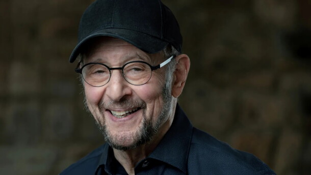 An older man in a ball cap, glasses and a dark blue button-down shirt smiles at the camera. 