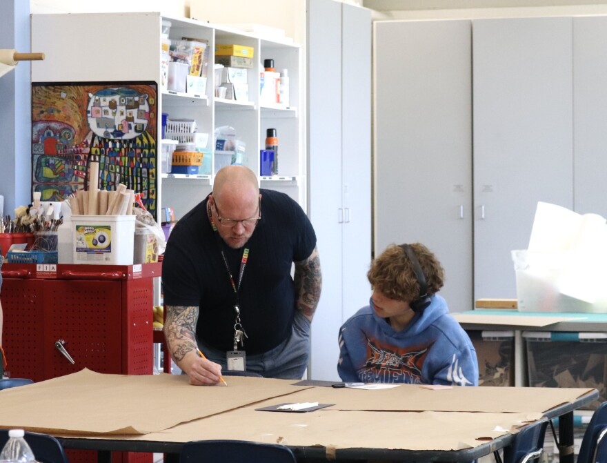 Matthew Osmon leans down to instruct Isaac Vasquez on the drawing of his design. Osmon holds a pencil and draws while his student tilts his head and observes. 