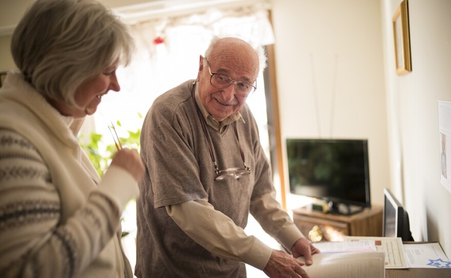An older gentleman stands in front of a desk and smiles at a woman assisting him. 