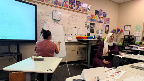A woman on the left is writing on a whiteboard. To her right, a woman wearing a hijab talks to her class.