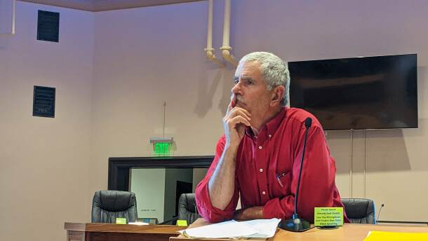 An old man stands at a podium inside a meeting room, he's looking off screen and pondering