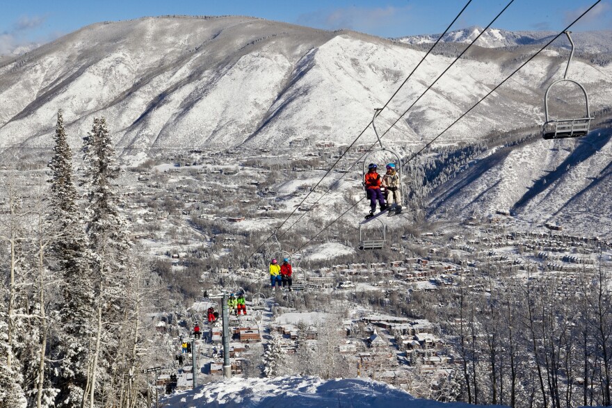Skiers and snowboarders ride up a ski lift with snowy hills and a town in the background.