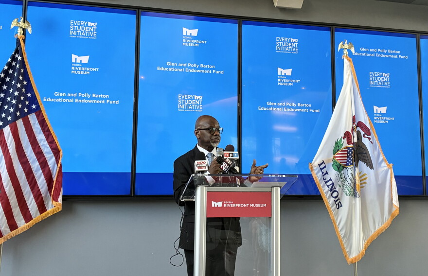 A man stands at a podium giving a speech, flanked by U.S. and Illinois flags. Behind him are blue screens displaying “Every Student Initiative,” Riverview Museum logos, and mention of the Glen and Polly Barton Educational Endowment Fund.