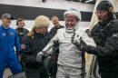 Man in white spacesuit and helmet smiles as he's helped by two people in dark, warm weather uniforms. Other people stand in the background.