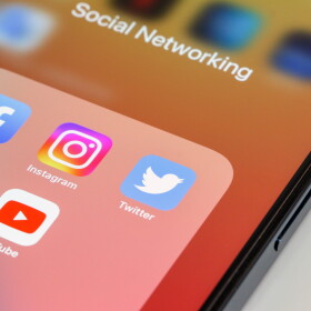 Close-up of popular social media app icons, Facebook, Instagram, Twitter, WhatsApp and YouTube, in that order, displayed on smartphone screen with orange wallpaper. Device is on white background.